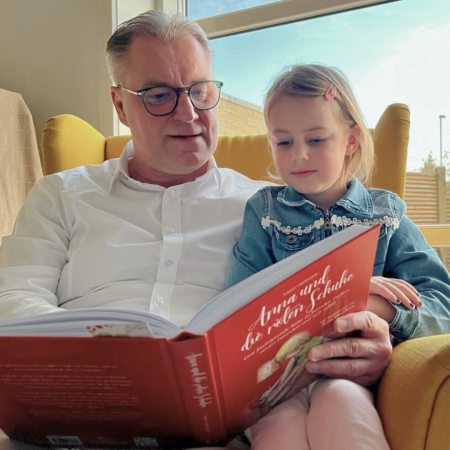 Foto von Mario Hartmann, der im Sessel sitzt mit einem Mädchen. Beide schauen in ein aufgeschlagenes Buch.